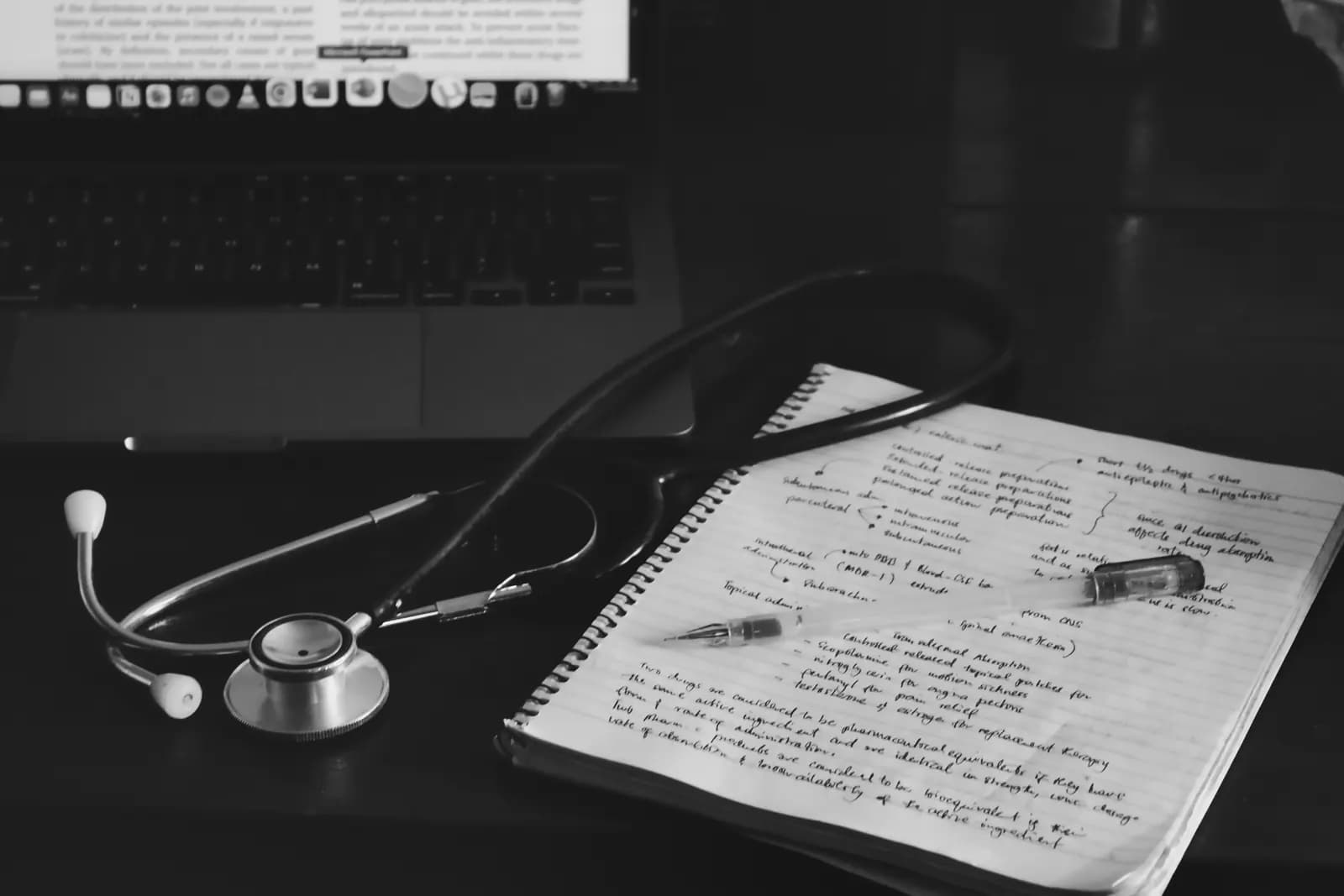 A stethoscope and handwritten pharmacology notes beside a laptop